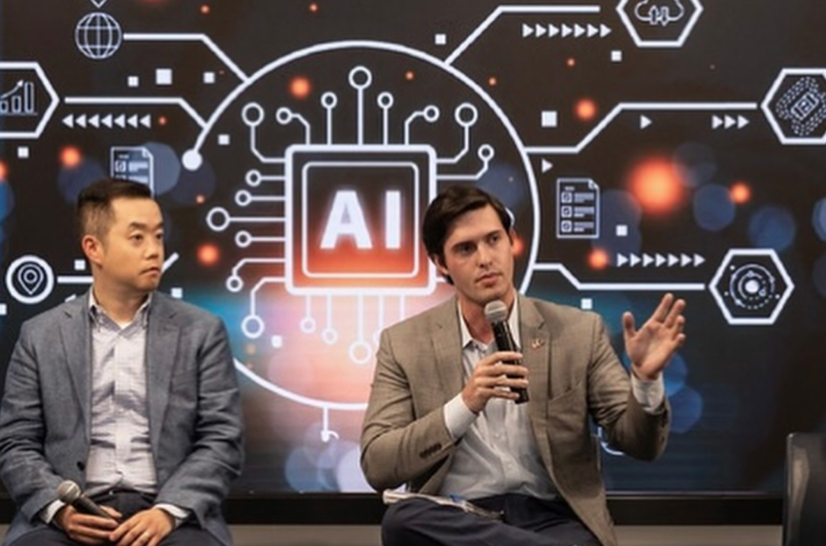 Colin Raby speaking on mike with a colleague seated next to him and an A.I. backdrop