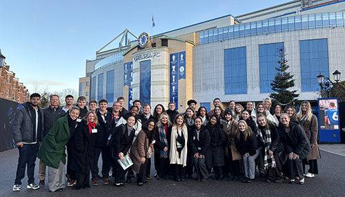 A large group of students including many LSU Tigers pose for a group photo outside the Chelsea Football Club headquarters in London. It is cold, the group is dressed warmly.